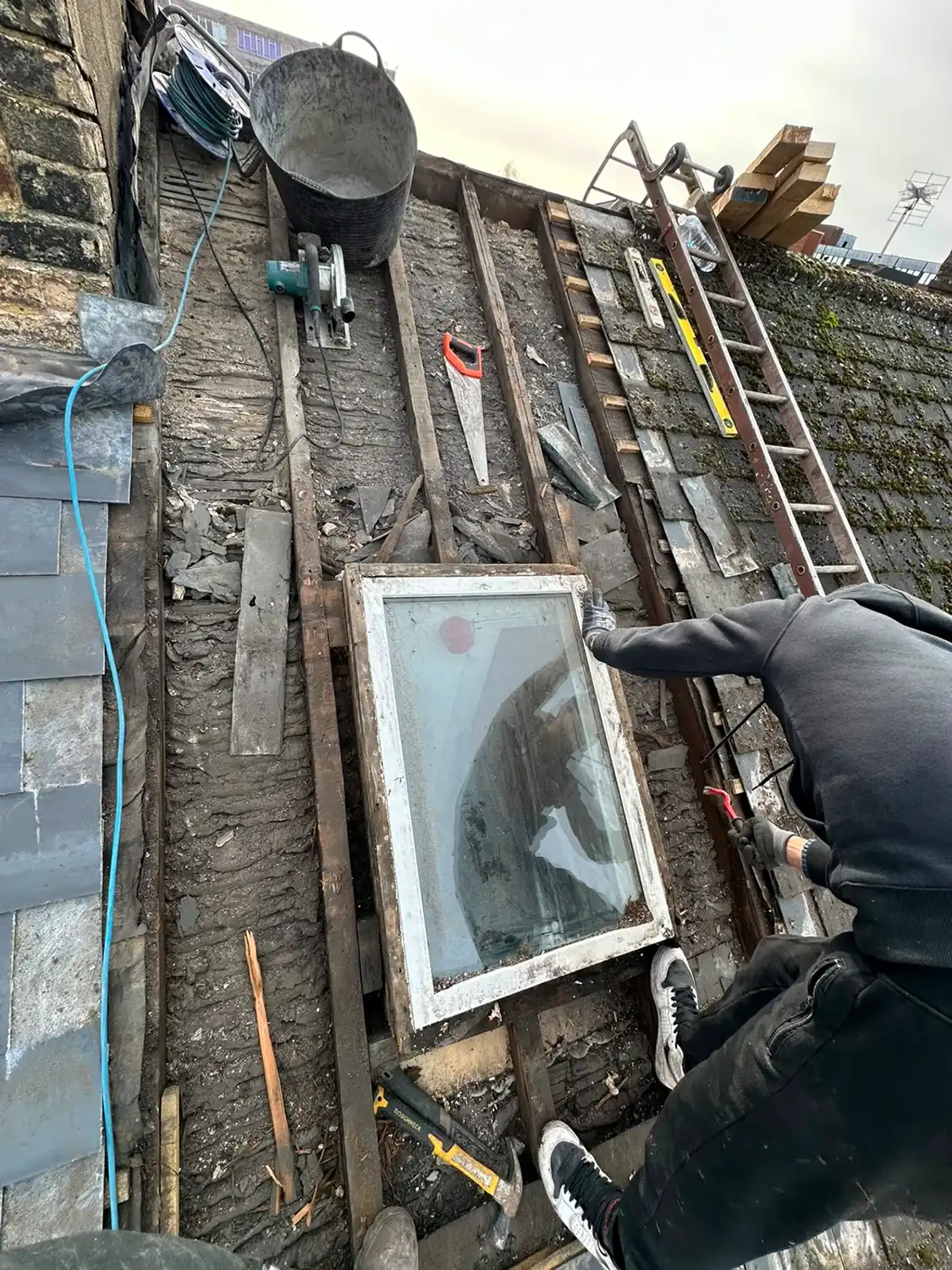 Person repairing a roof with exposed wooden beams, tools, and a window frame ready to be installed.