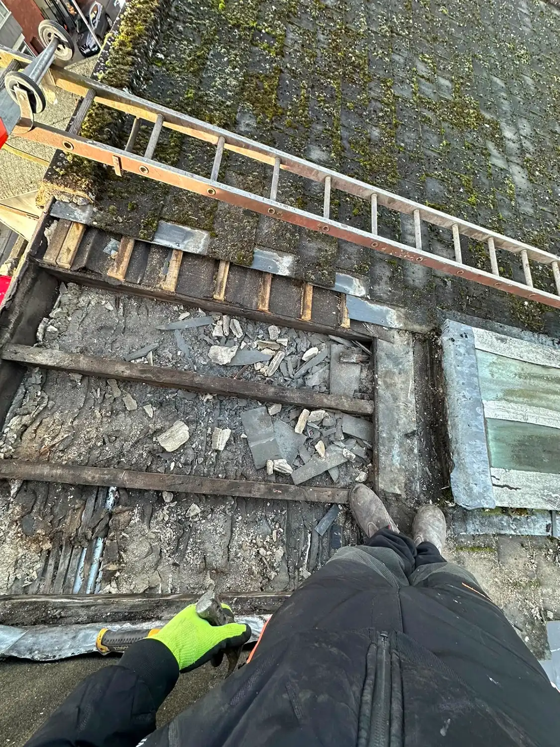 View from roof showing a worker's boots and gloved hand near a partially removed mossy roof section.