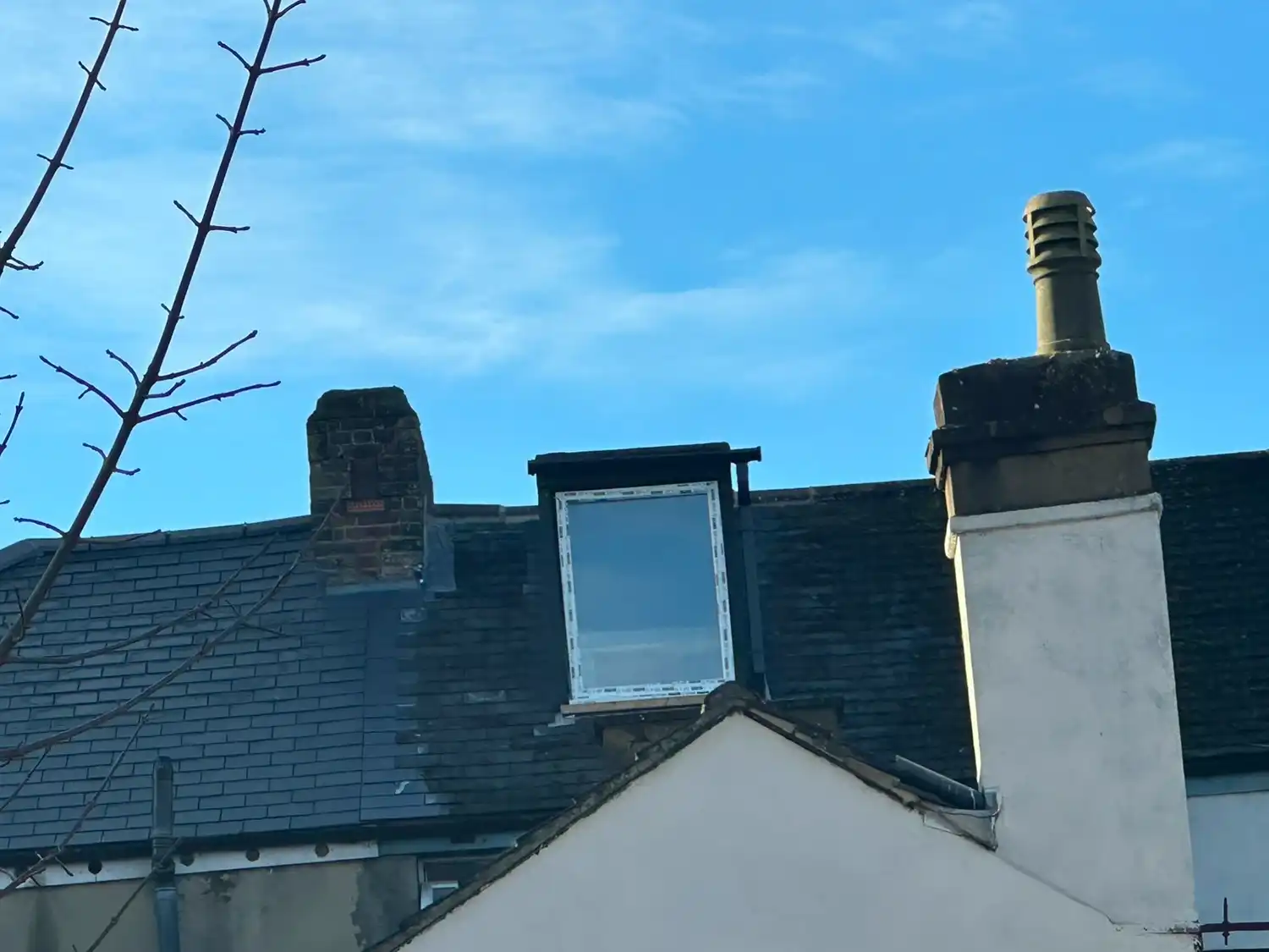 Rooftop with chimney, a closed dormer window, and a leafless tree branch under a blue sky.