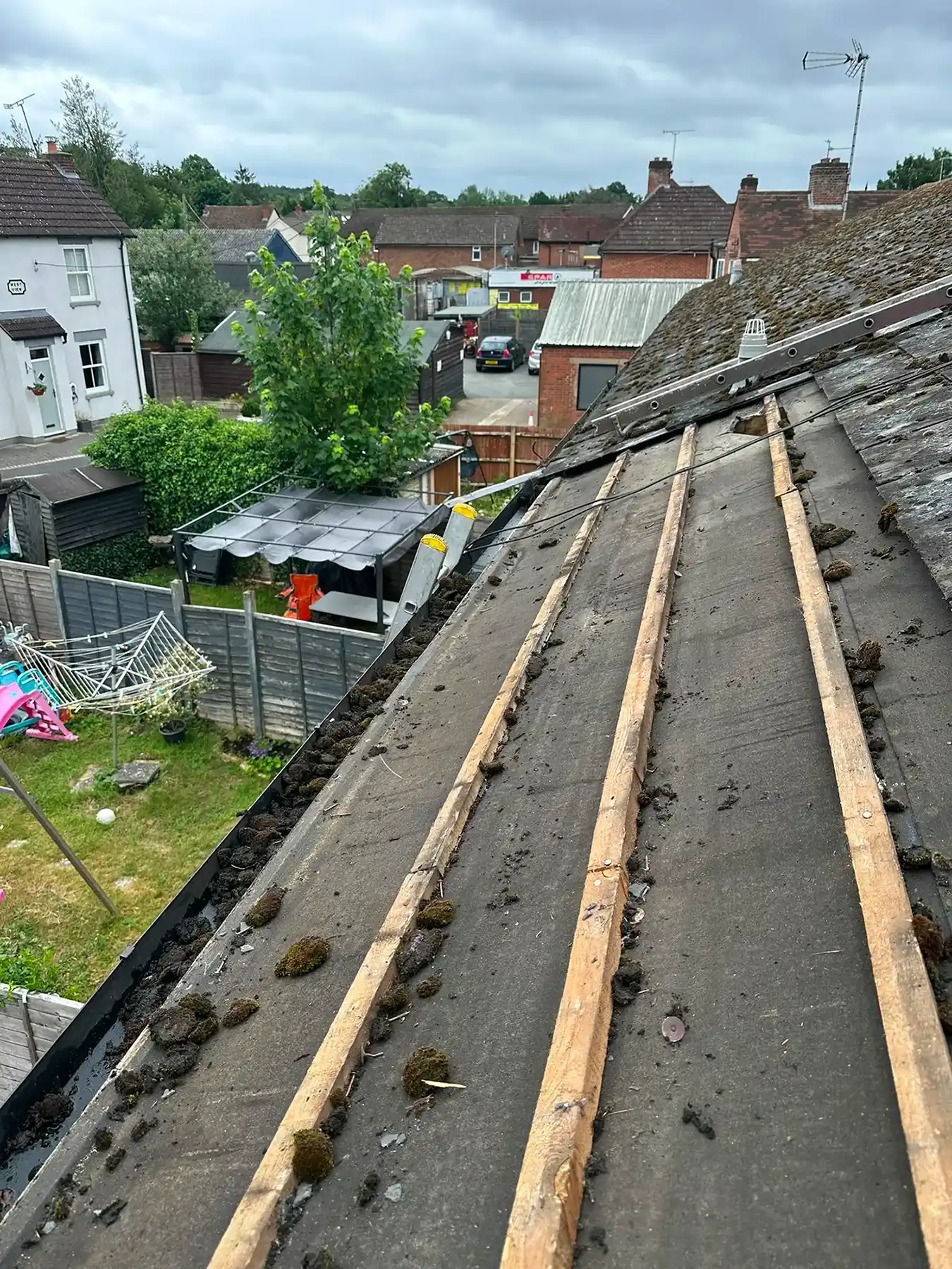Roof with wooden battens and patches of moss, overlooking a backyard with garden and houses beyond.
