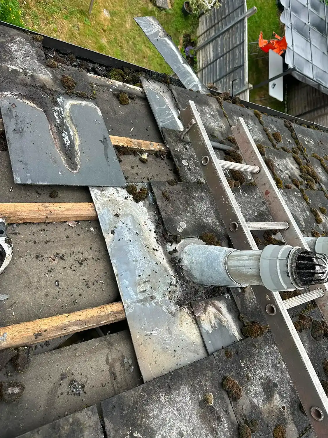 Close-up of a mossy roof with exposed wood beams, metal flashing, and a metal chimney pipe.