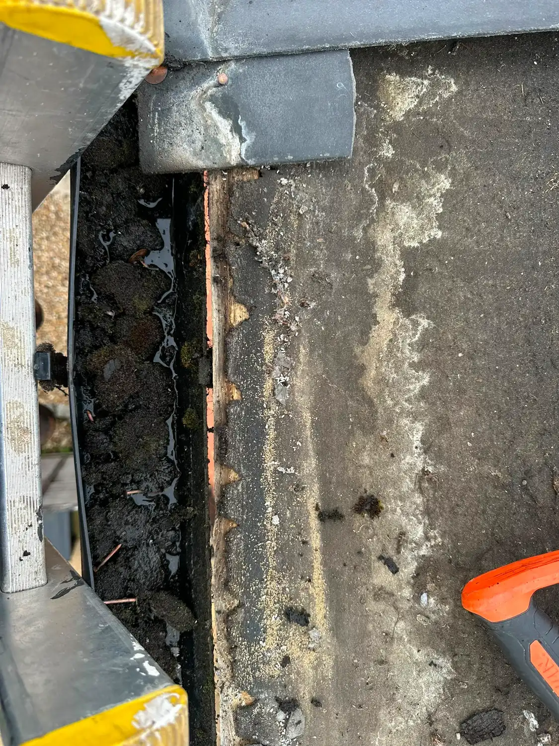 Close-up of a dirty roof gutter with accumulated debris and part of an orange tool handle.
