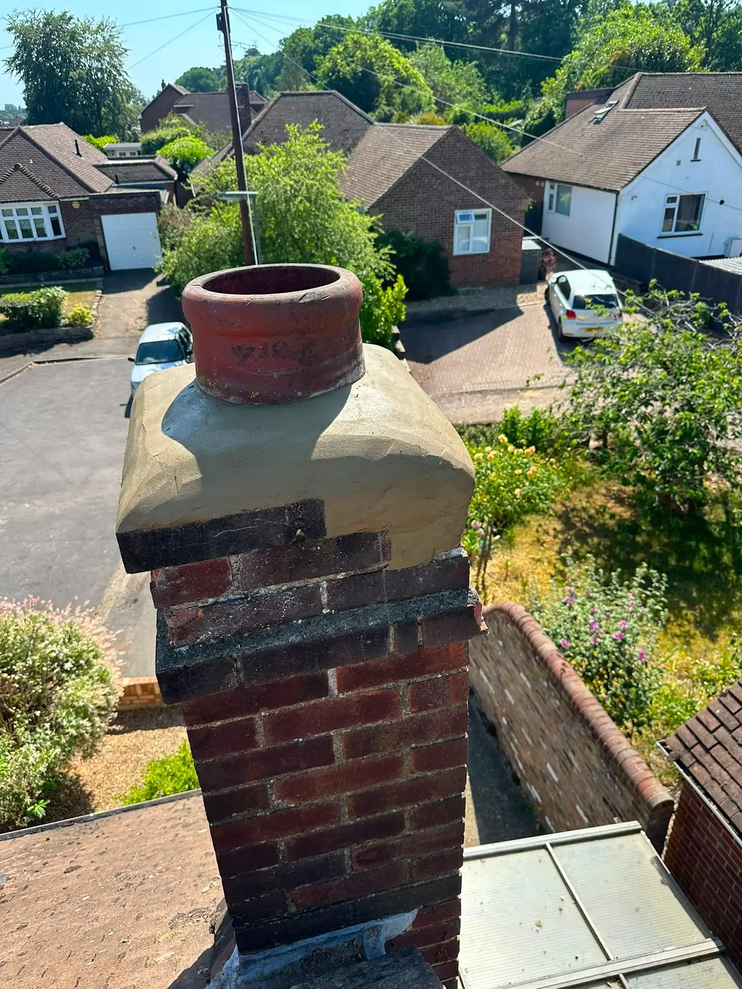 Brick chimney with clay pot on a house roof, overlooking neighboring houses and greenery on a sunny day.