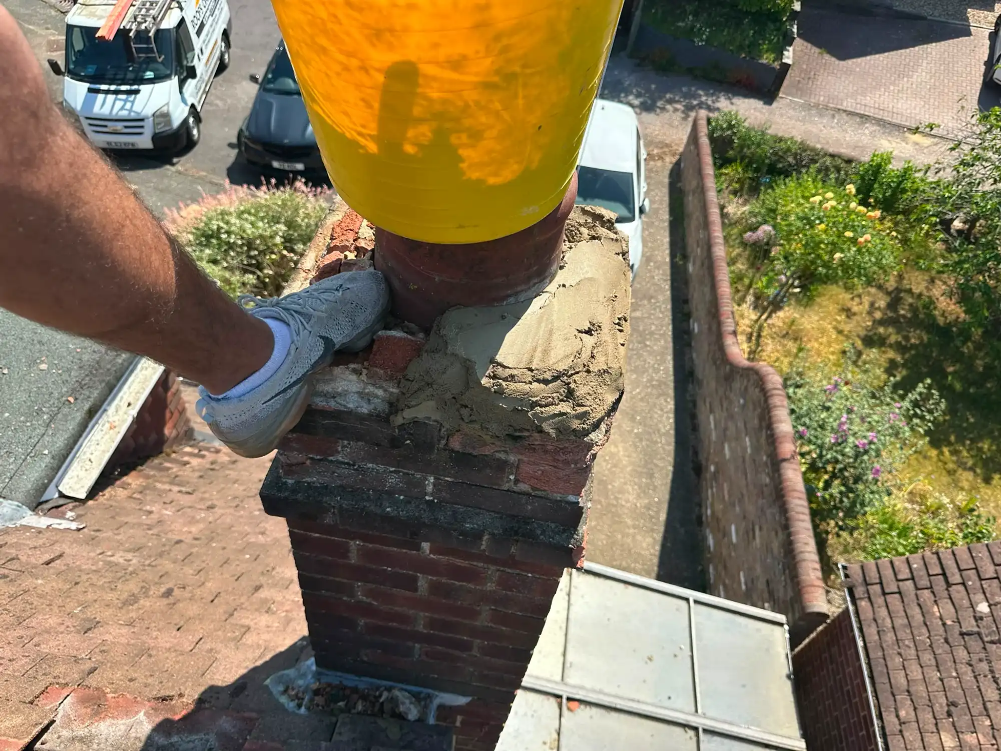 Person's leg and foot in gray shoe resting on a brick chimney topped with cement and a yellow bucket.