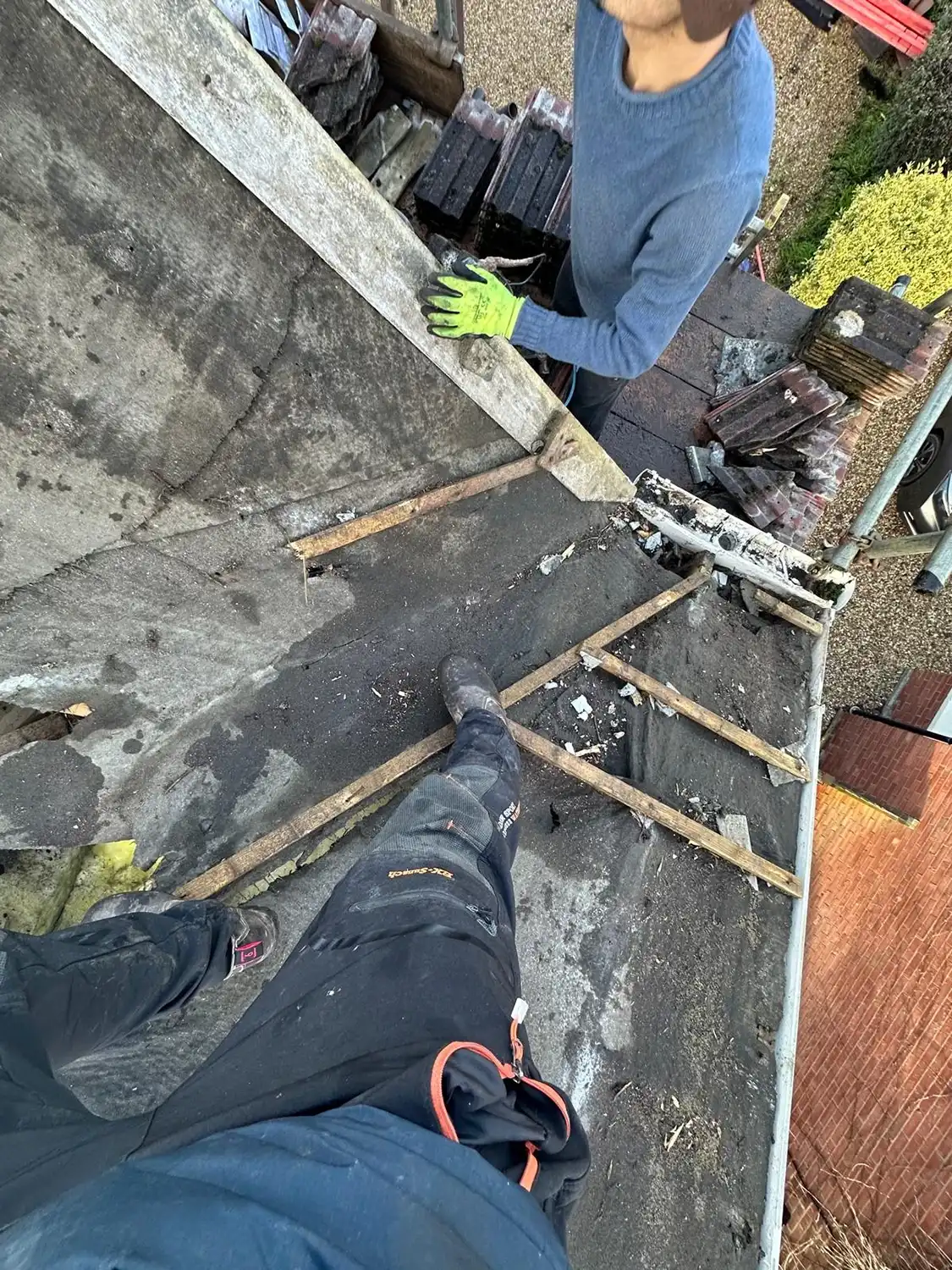 Person in black pants standing on roof next to another person in blue sweater and green gloves.
