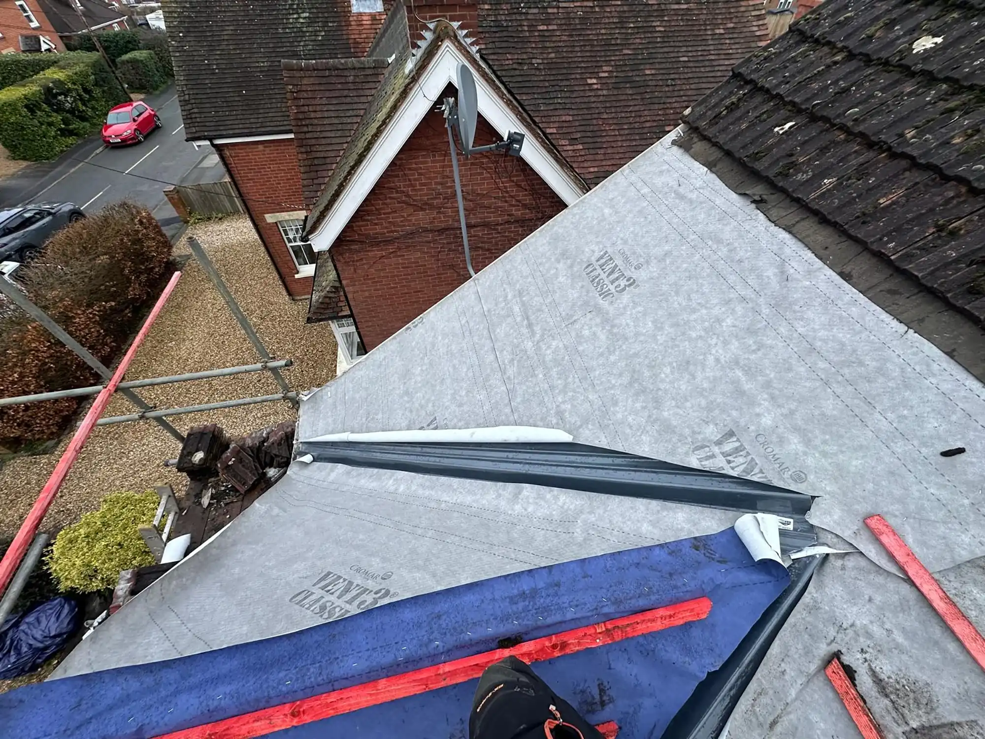 View over a roof under construction with protective membrane and scaffolding, and nearby brick house with satellite dish.
