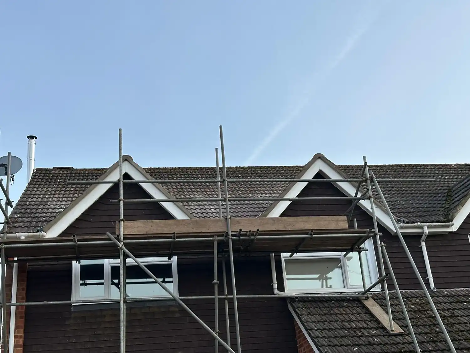 Scaffolding set up in front of a dark brown house with steep tiled roof and two attic windows.