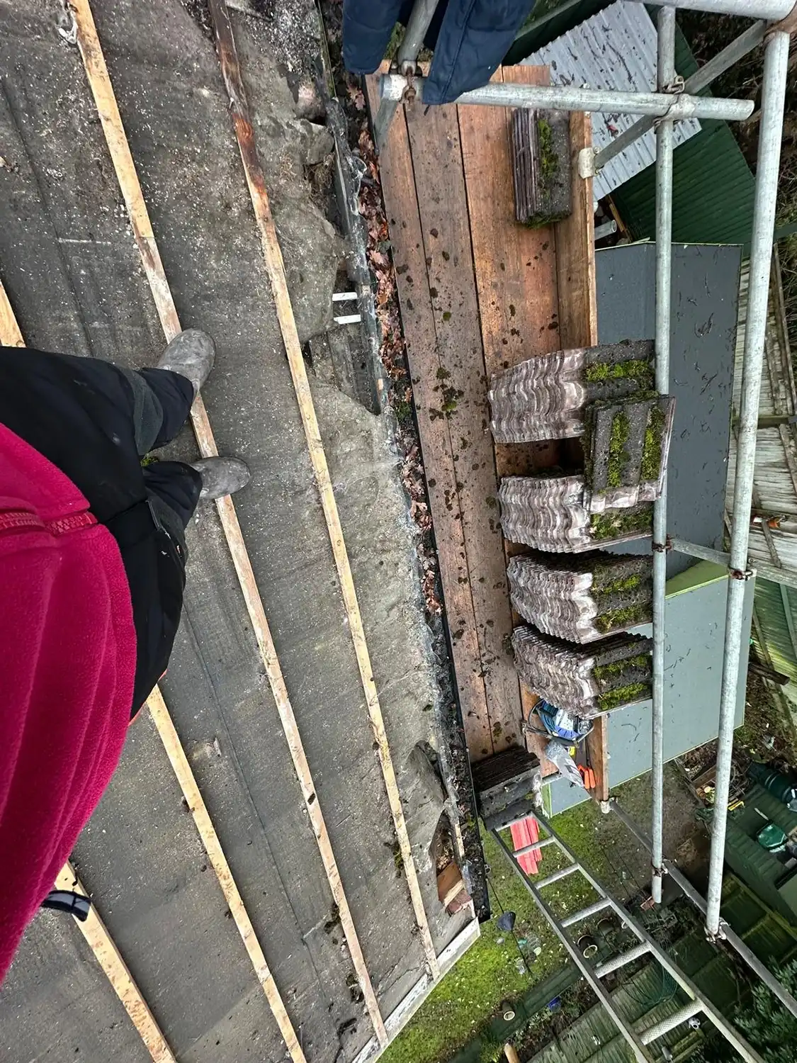 View of a person standing on a roof under repair with wooden battens and stacks of mossy tiles on scaffolding.
