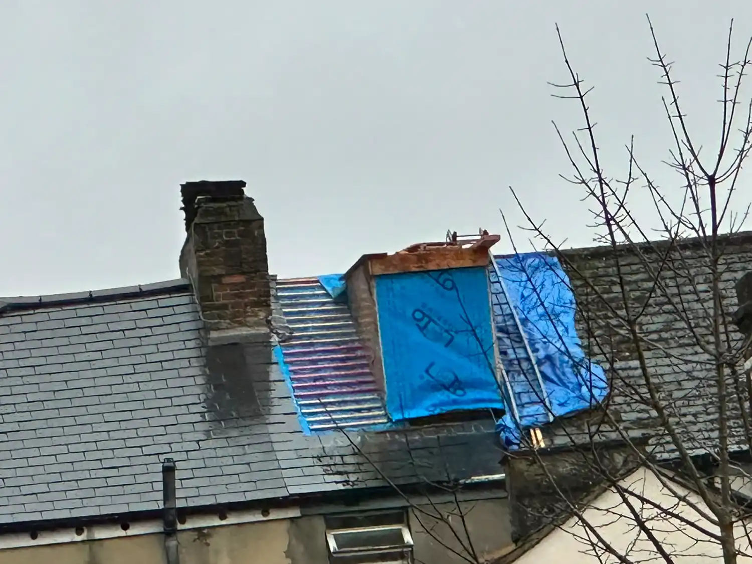 Roof with missing tiles covered partly by blue tarps and a chimney on a cloudy day.