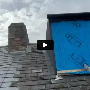 Roof section with slate tiles, a chimney, and blue construction sheathing on a dormer under a cloudy sky.